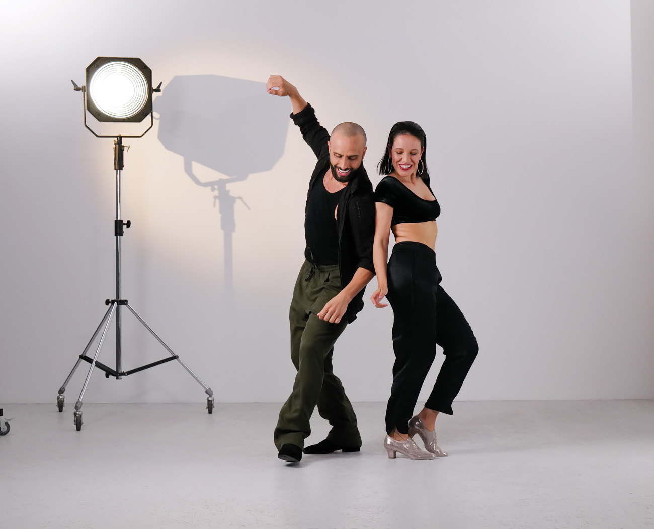 Bachata-3 Dynamische Bachatamoves von jungem Tanzpaar in lässigen dunklen Outfits mit Tanzschuhen von Werner Kern in silber und schwarz vor weißer Studiowand mit Lichtstrahler im Hintergrund