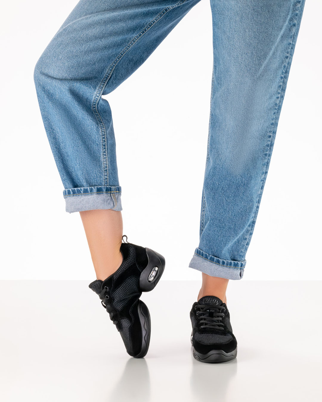 Modell in Jeans trägt schwarze Tanzseaker mit geteilter Sohle und biegt den rechten Fuß über den Spann um die Biegsamkeit des Schuhs darzustellen